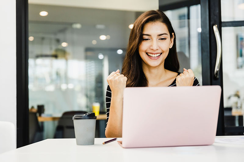 A woman sitting at a desk, smiling and clenching her fists in excitement while looking at her laptop.