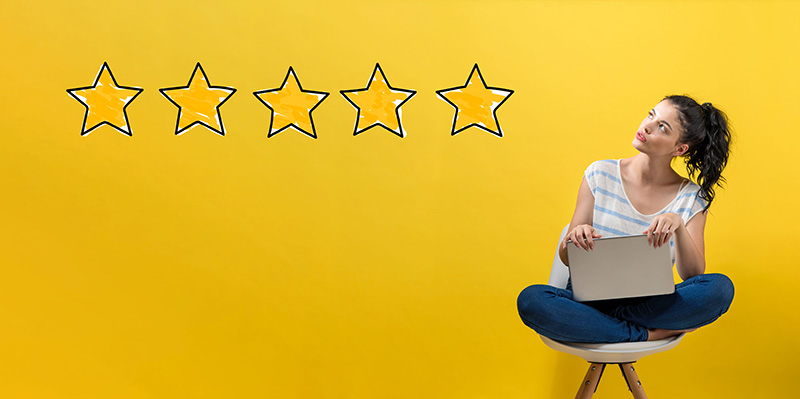 An image of a woman sitting crossed-legged on a stool, looking at a row of five giant yellow stars on her left, against a yellow background, representing a review.