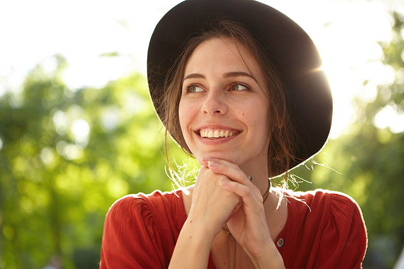 Smiling woman wearing a hat outdoors, enjoying a sunny day with a cheerful expression.